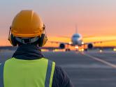 Airport ground crew directs aircraft at sunset wearing a safety vest and hardhat.